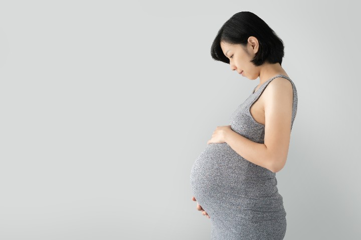 Pregnant Asian woman standing and touching big belly with her hands on gray Isolated background