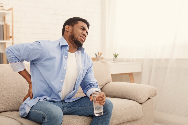 Young african-american man with back pain, pressing on hip with painful expression, sitting on sofa at home with glass of water, copy space