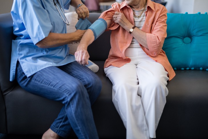 Low section of doctor checking senior woman blood pressure while sitting on sofa at home