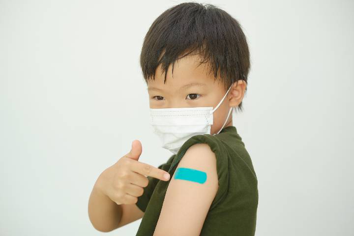 Doubtful child after getting a vaccine. Little kid wearing medical mask showing his arm with bandage after receiving vaccination, COVID 19 vaccine for children and youth, Back to school concept